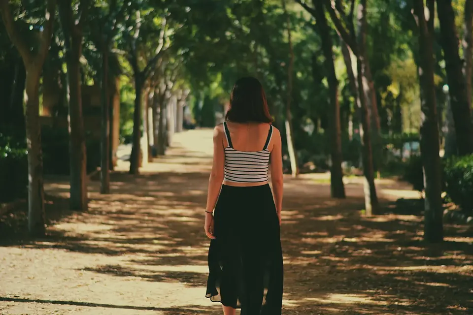 A woman walking alone along a wide, tree-lined path in a city park